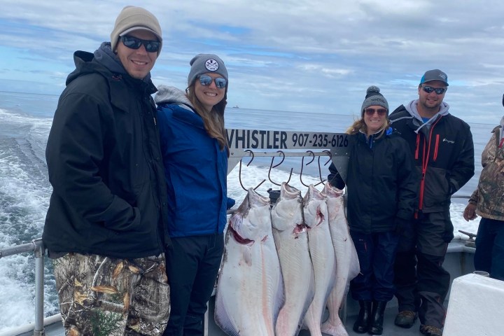 Group of five people on a boat posing with five large caught fish hanging on hooks.
