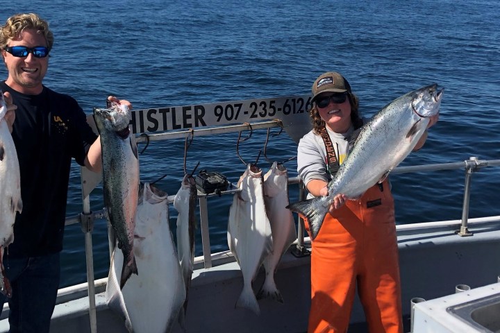 Two people on a boat holding large fish, with additional fish hanging in the background.