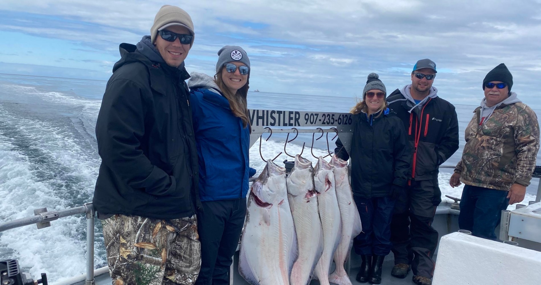 Five people on a boat posing with large fish hanging on hooks under a cloudy sky.