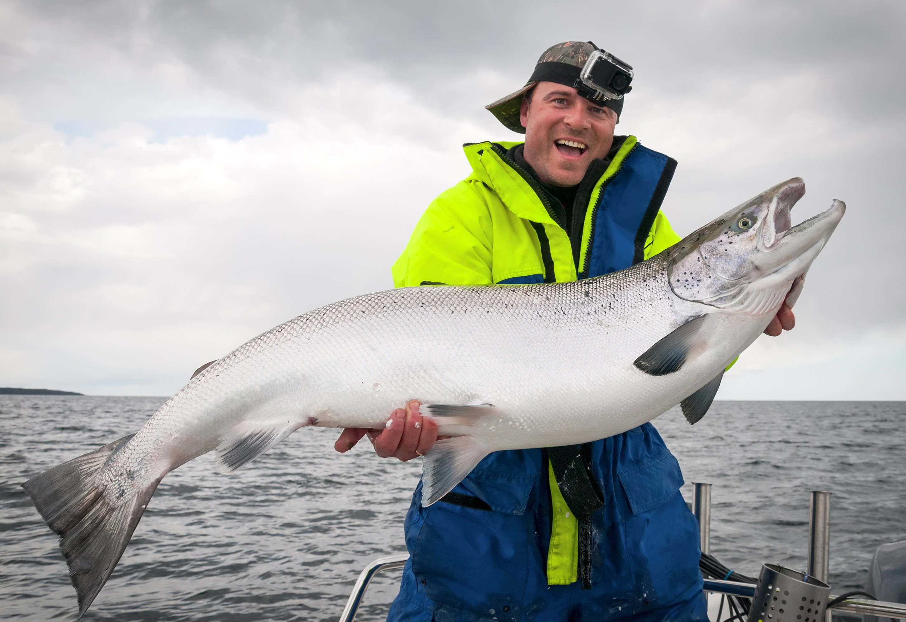 Person in bright jacket holding a large fish on a boat with cloudy sky.