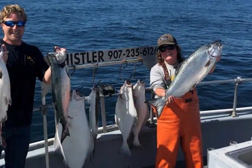 Two people on a boat holding large fish with the sea in the background.