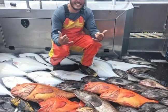 Smiling fisherman in orange overalls with large catch of fish on a boat deck.