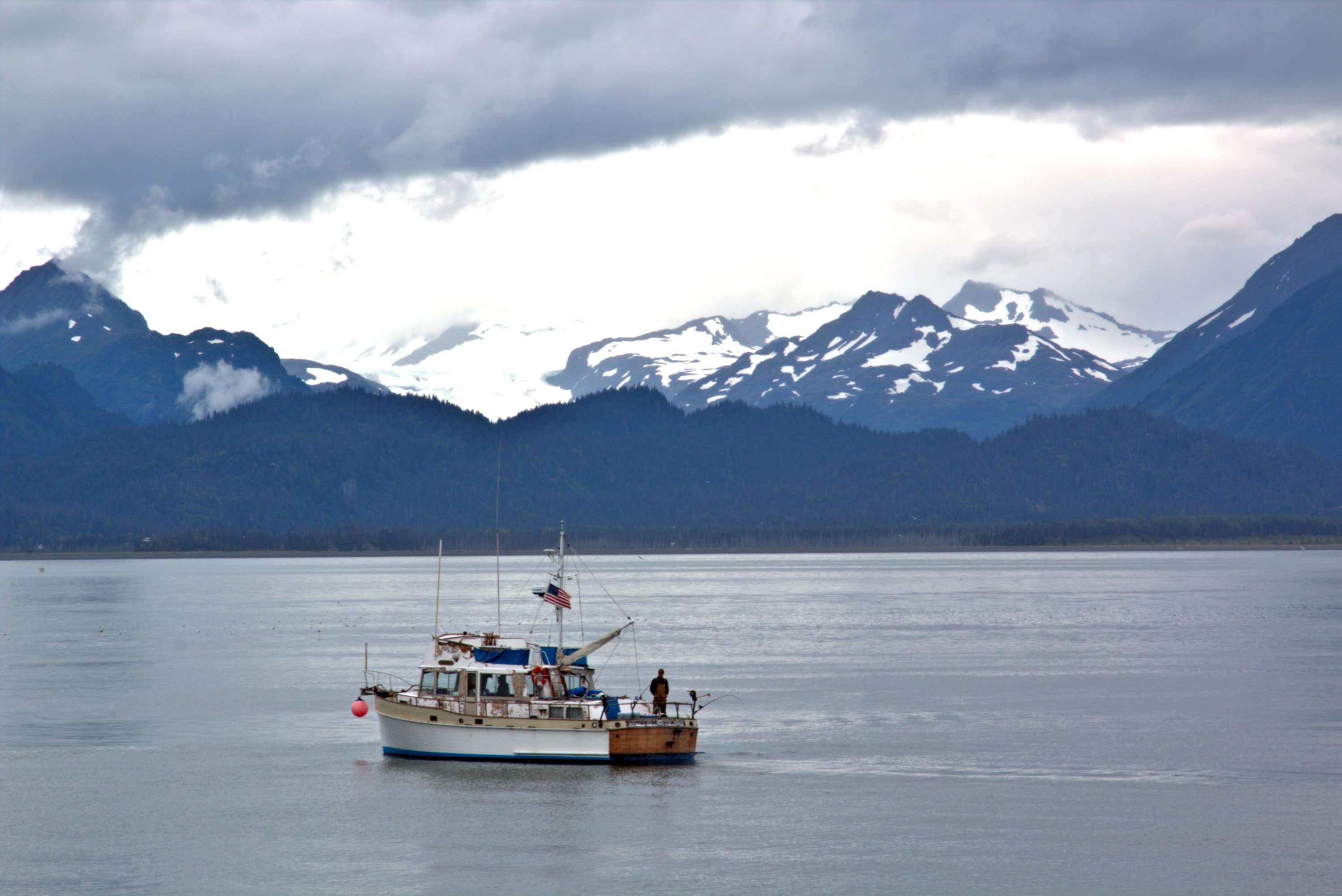 Boat on calm water with snow-capped mountains and cloudy sky in background.